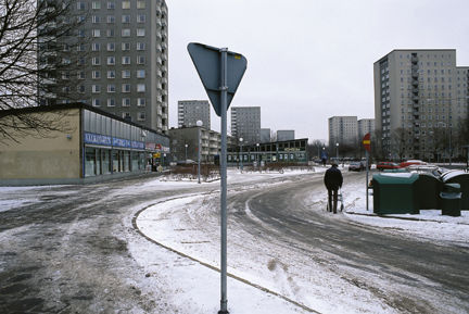 En väg med tunt lager snö, låga och höga hus i bakgrunden.