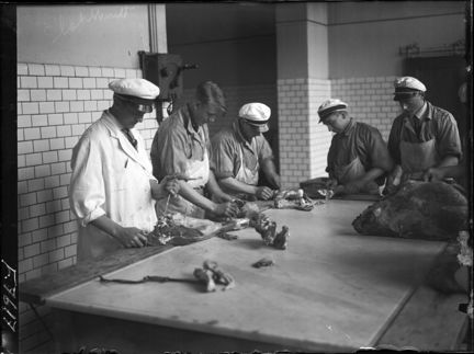 Fem män står vid ett bord och stycker kött. 