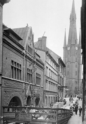 I fonden syns en kyrka med ett högt och spetsigt torn. Till vänster i bild ser vi en rad låga stenhus. Närmast i bild syns en nedfart till en tunnel. På ett staket syns skyltar som säger att det är förbjudet att parkera i transporttunneln och att obehöriga ej äger tillträde till arbetsplatsen. 