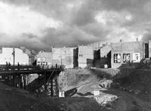 Vy över rivna kv. Stigbygeln österut. Mäster Samuelsgatan går på en provisorisk bro. Här ska tunnelbanan anläggas och blivande Sveavägen dras fram. Tv. kv. Putten och t.h. kv. Hästryggen
