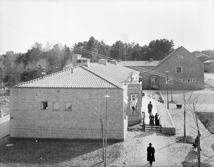 Ett mindre torg omges av en låg och en lite högre tegelbyggnad. Torget är belagt med grus och har nyplanterade träd och rabatter. Några människor är på väg till och från den livsmedelsbutik som finns i den låga byggnaden.  