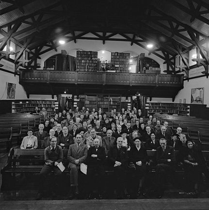 Bilden är tagen från en scen, rakt framför en sittande publik som tittar in i kameran. De sitter i en stor sal som har en läktare på den bortre väggen. I taket syns stora träbjälkar. I rummet och på läktaren finns många bokhyllor fulla med böcker. 