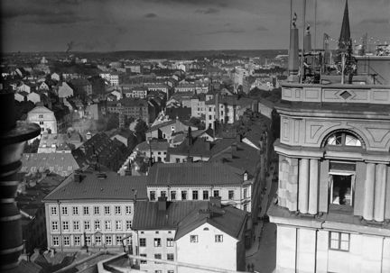 En vy över staden tagen högt uppifrån ett torn, det södra Kungstornet. Närmast i bild syns toppen av det norra Kungstornet. På toppen av det tornet syns en liten uteservering med upphängd belysning. I balkgrunden breder staden ut sig med mängder av stenhus i flera våningar. 