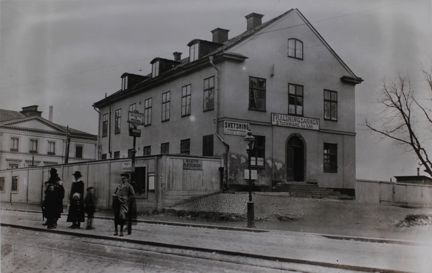 Ett stenhus i två våningar med takkupor. På fasaden sitter skyltar som visar att det är Frälsningsarméns hus och att det ligger en svetsverkstad i bottenvåningen eller i närheten. Utanför huset finns en spårvagnshållplats där några vuxna och barn står och väntar.