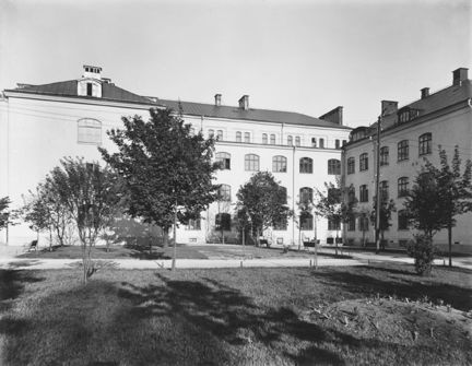 Ljus byggnad med gräsmatta och träd i förgrunden.