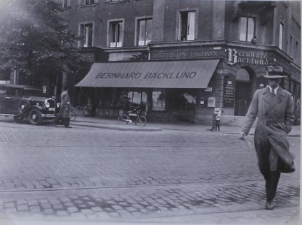 I förgrunden en man i hatt och rock. I bakgrunden Bäcklunds Herrekipering. På fasaden står "Herrkonfektion" och "Bernhard Bäcklund". På en stor markis står "Bernhard Bäcklund".