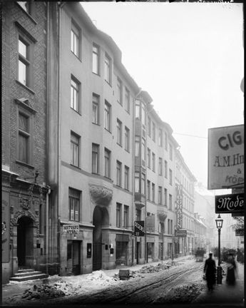 Dominerande i bilden är ett stort, ljust putsat stenhus i så kallad jugendstil. På gatan är det snömodd och människor promenerar längs trottoarerna. Butiksskyltar hänger ut från fasaderna.